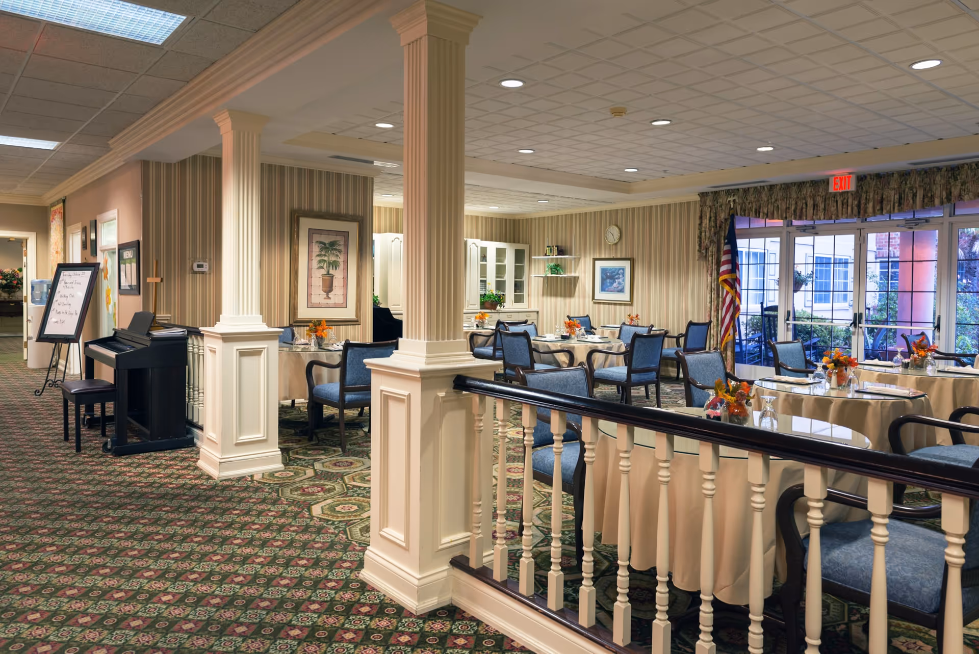 Interior view of a dining room in a senior living facility with round tables covered in beige tablecloths, each set with chairs and small floral centerpieces. The room features patterned carpet, striped wallpaper, decorative columns, and large windows with an exit door leading outside. An American flag is positioned near the door, and a piano with a bench is visible along the left side of the room.