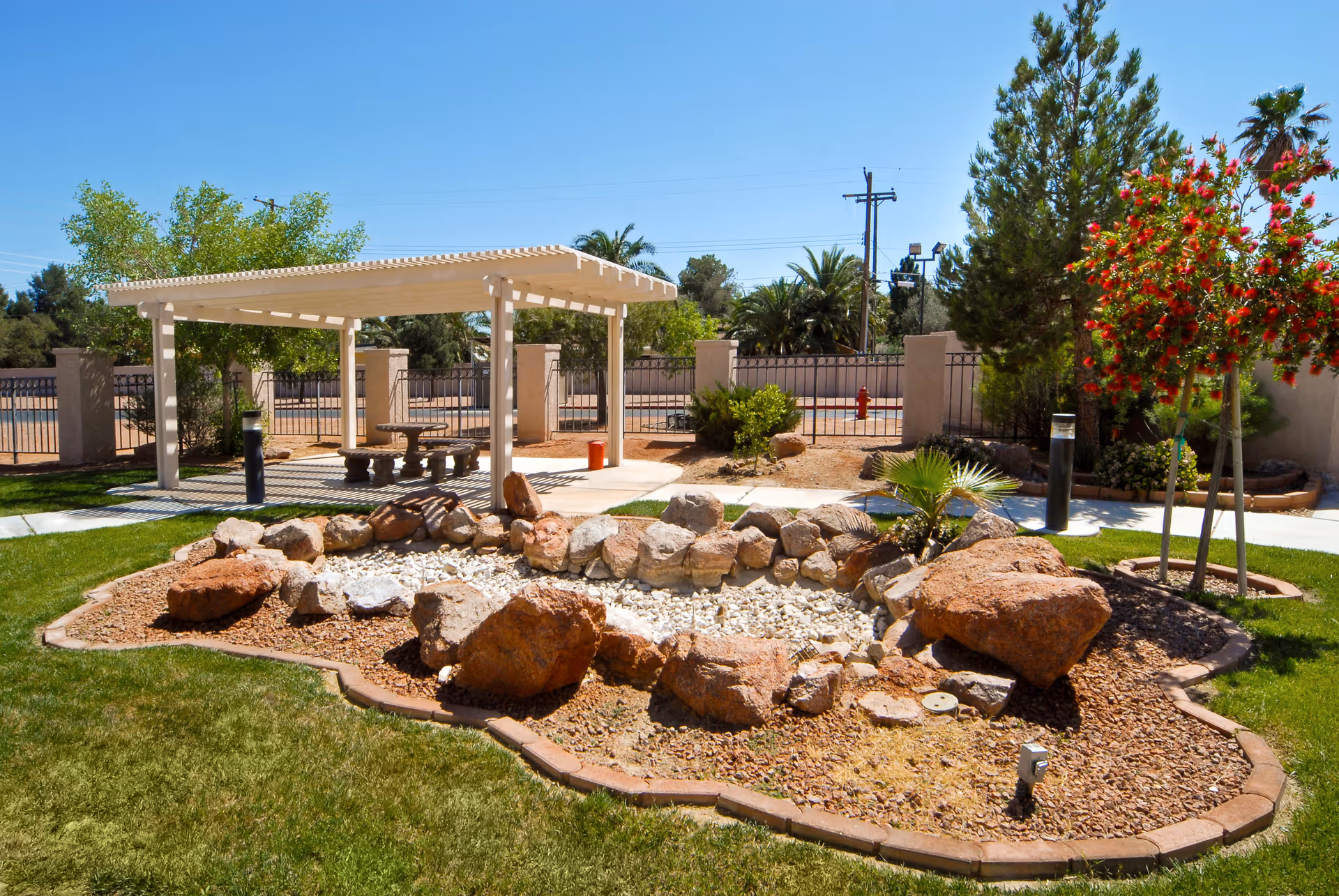 Outdoor garden area with a dry rock pond surrounded by large rocks and small plants. There is a white pergola with a picnic table and benches underneath it. The area is fenced with a metal fence and there are trees and shrubs around the garden. The sky is clear and blue.