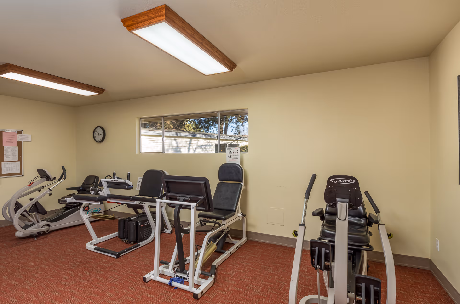 A small exercise room with various fitness equipment including a recumbent bike, a rowing machine, and a leg press machine. The room has beige walls, a red carpeted floor, a window letting in natural light, and fluorescent ceiling lights.