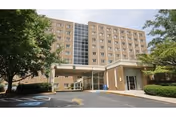 Exterior view of a multi-story senior living facility building with a covered entrance driveway. The building has multiple windows and is surrounded by trees and landscaping. The sky is clear with some clouds.