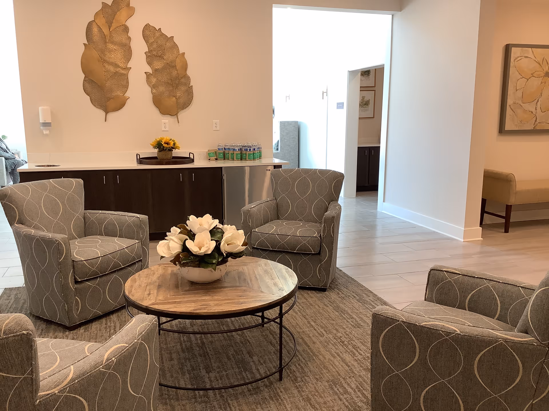 A bright lounge with four patterned armchairs around a round wooden coffee table topped with a flower arrangement, and cabinetry and wall decorations in the background.