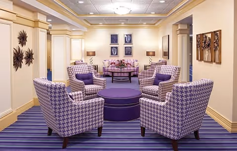 A cozy sitting area with four purple and white houndstooth patterned armchairs arranged around a round purple ottoman. The room has a striped purple carpet, cream-colored walls with decorative molding, and framed artwork on the walls. In the background, there is a round table with a floral centerpiece and two table lamps on either side.
