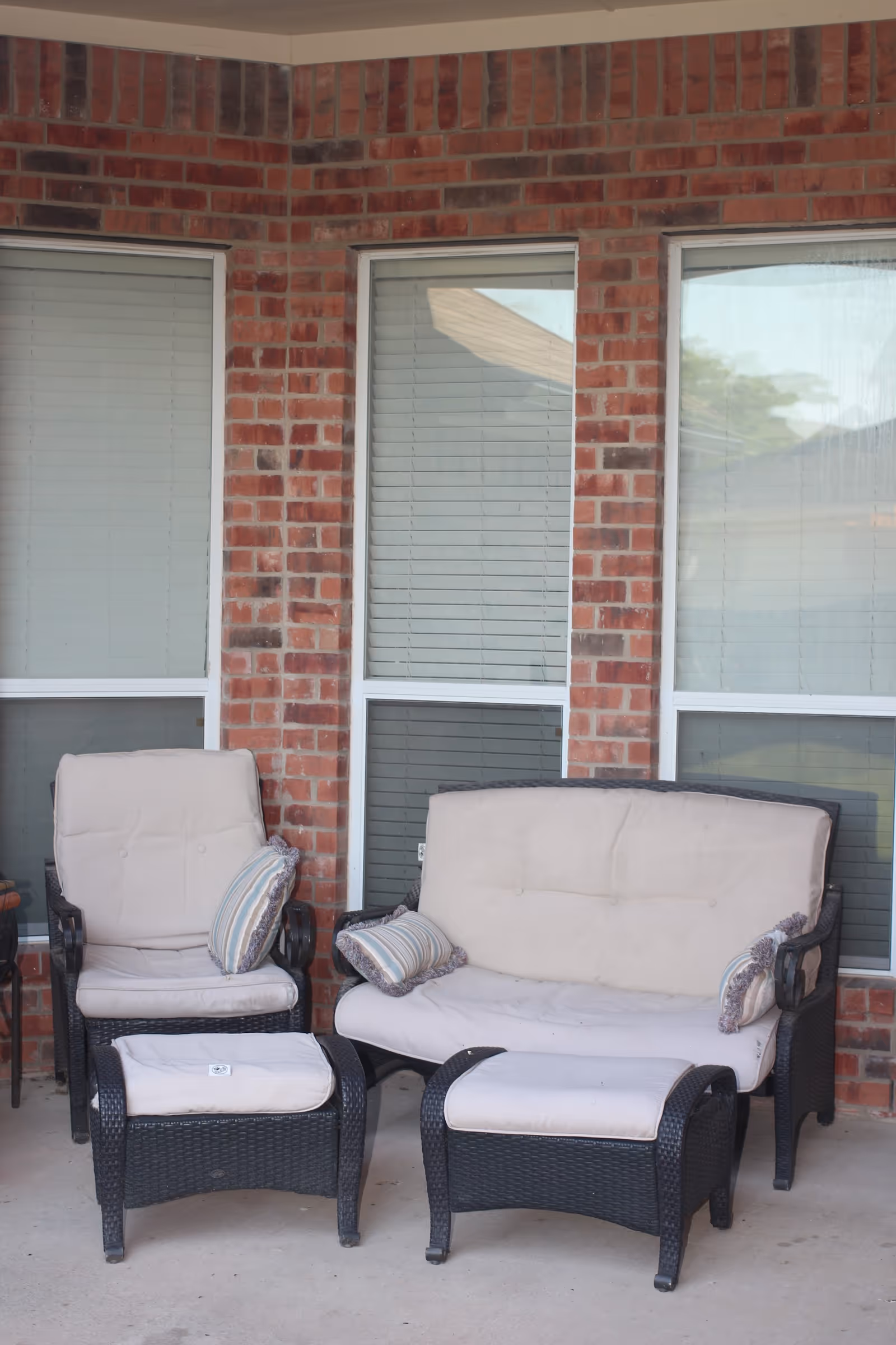 Outdoor patio area with a cushioned armchair, a cushioned loveseat, and two cushioned ottomans in front of a brick wall with three windows.