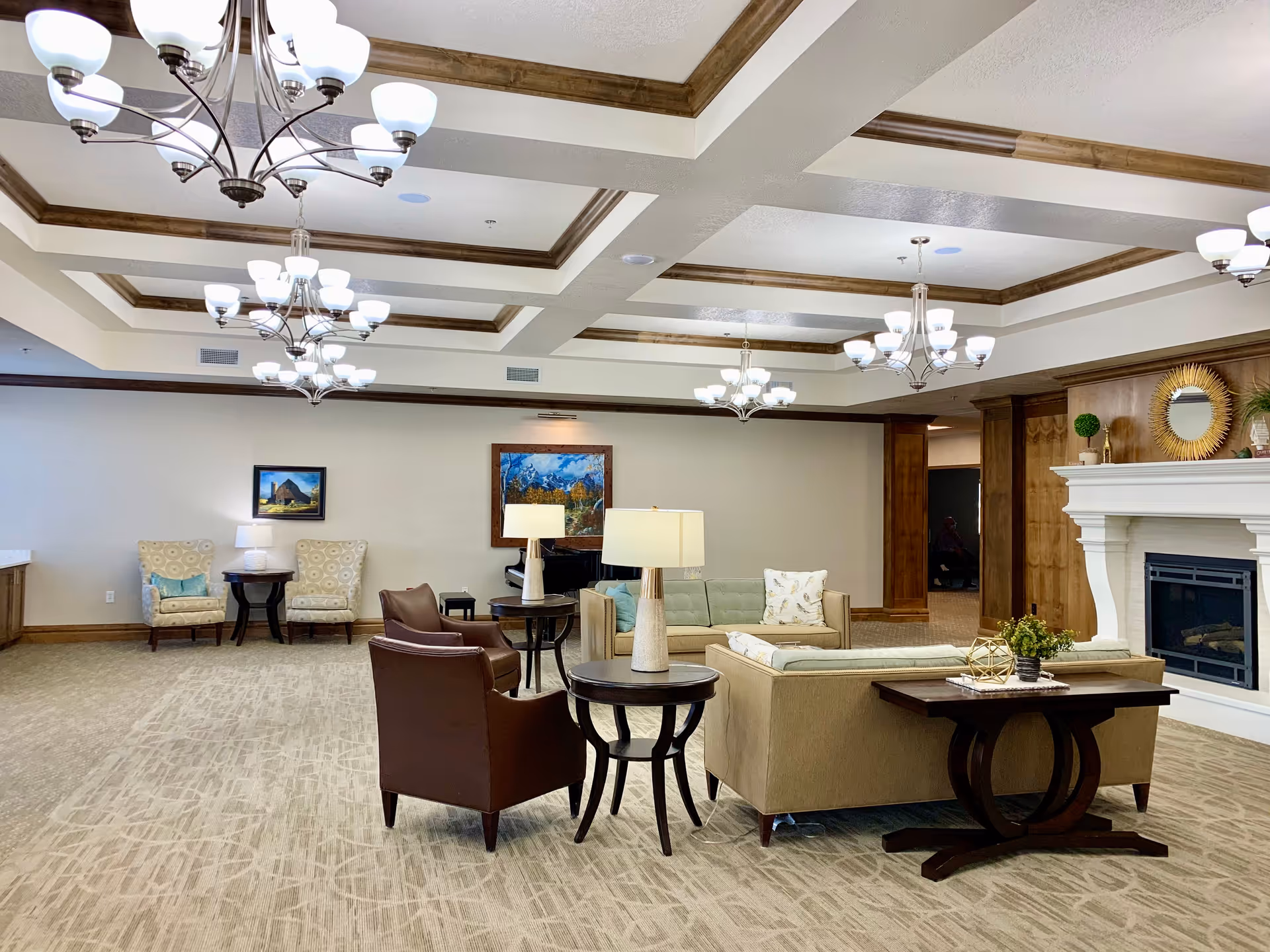 A spacious and well-lit living room area in an assisted living facility featuring beige carpeting, multiple chandeliers with white lampshades, a white fireplace with decorative items on the mantel, a beige sectional sofa with pillows, two brown leather armchairs, side tables with lamps, and framed artwork on the walls.