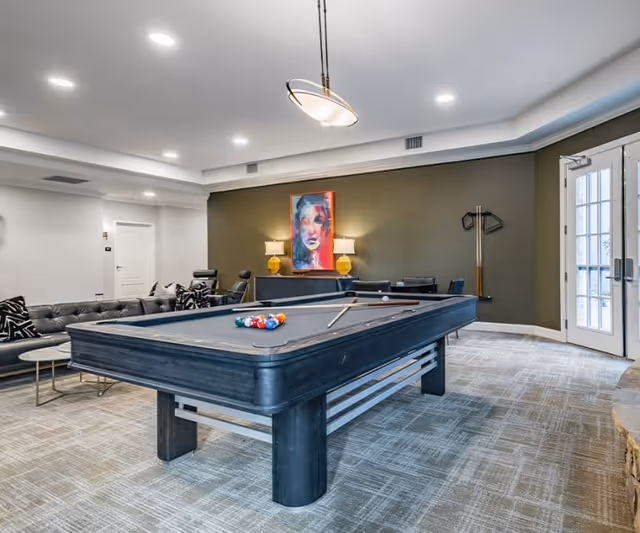 A modern game room featuring a black pool table with billiard balls and cues arranged on it. Behind the pool table is a seating area with a black leather sectional sofa and a small round table. The room has a green accent wall with a colorful abstract portrait painting, two yellow lamps on a console table, and a coat rack. There is a door with glass panels on the right side, and the room is well-lit with recessed ceiling lights and a hanging light fixture above the pool table.