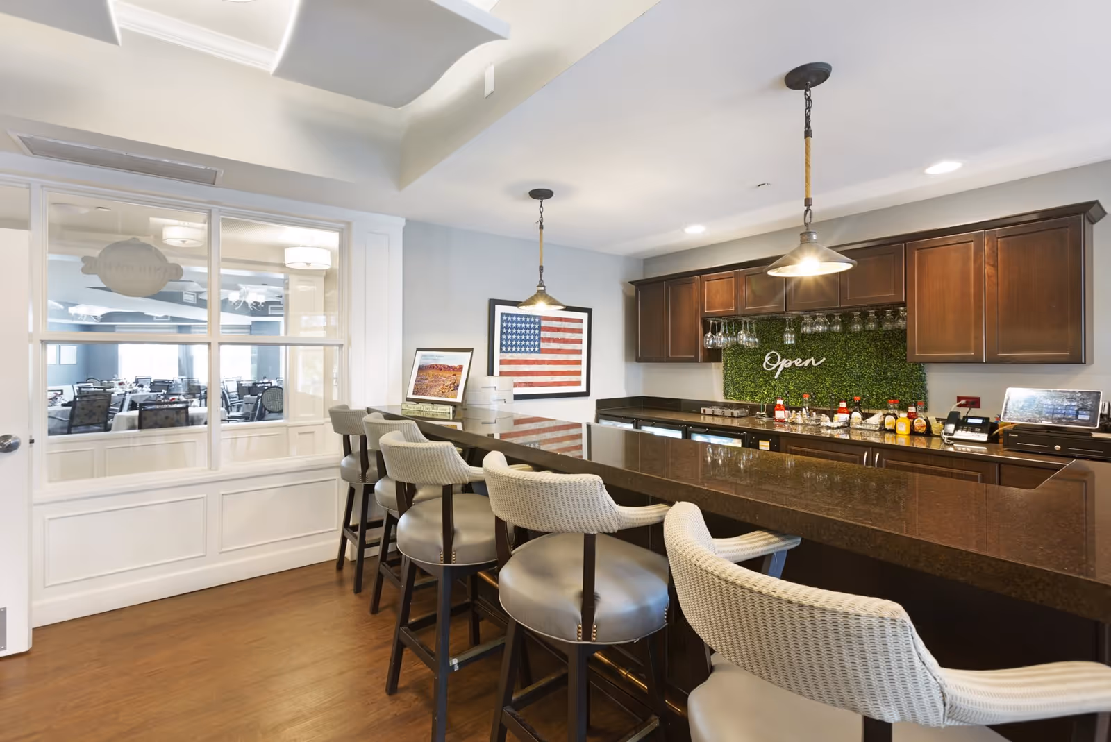 A modern bar area with a long dark countertop and several cushioned bar stools. Behind the bar are wooden cabinets, hanging wine glasses, and a green wall with the word 'Open' illuminated. There is an American flag artwork on the wall and a window showing a dining area with tables and chairs.