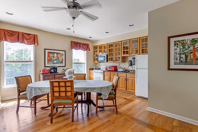 A bright kitchen and dining area with wooden cabinets, a white refrigerator, microwave, toaster, and coffee maker. A round dining table with a patterned tablecloth is surrounded by four wooden chairs. Two large windows with red valances let in natural light. The room has hardwood floors, a ceiling fan, and framed artwork on the walls.