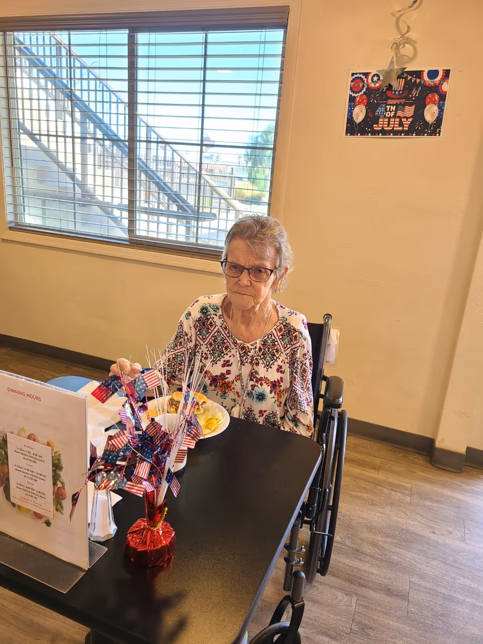An elderly woman in a wheelchair sitting at a dining table with a plate of food in front of her. The table has a small patriotic centerpiece with miniature American flags. Behind her is a window with blinds and a wall decorated with a 'Happy 4th of July' sign.