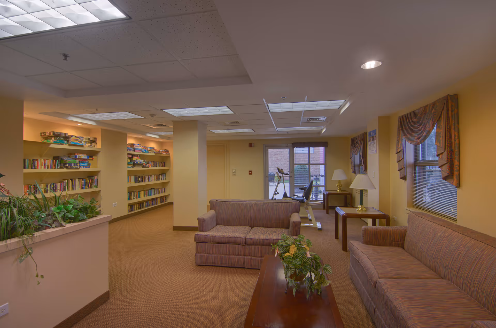 A cozy common area in a senior living facility featuring two striped sofas, a wooden coffee table with a plant, side tables with lamps, and large windows with patterned valances. In the background, there are bookshelves filled with books and board games, and exercise equipment near a glass door leading outside.