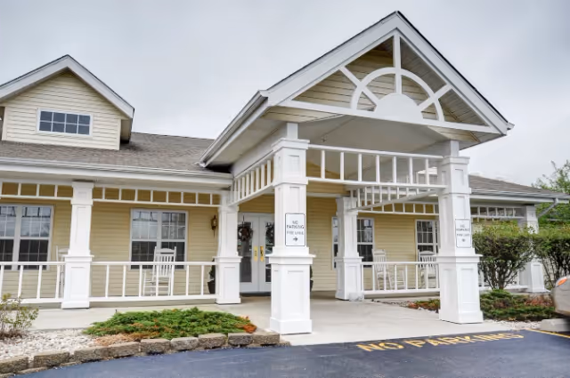 Exterior view of a senior living facility entrance with a covered porch supported by white columns. The building has beige siding, multiple windows, and a paved driveway with 'NO PARKING FIRE LANE' painted on it. There are white rocking chairs on the porch and some landscaping with bushes and small plants.