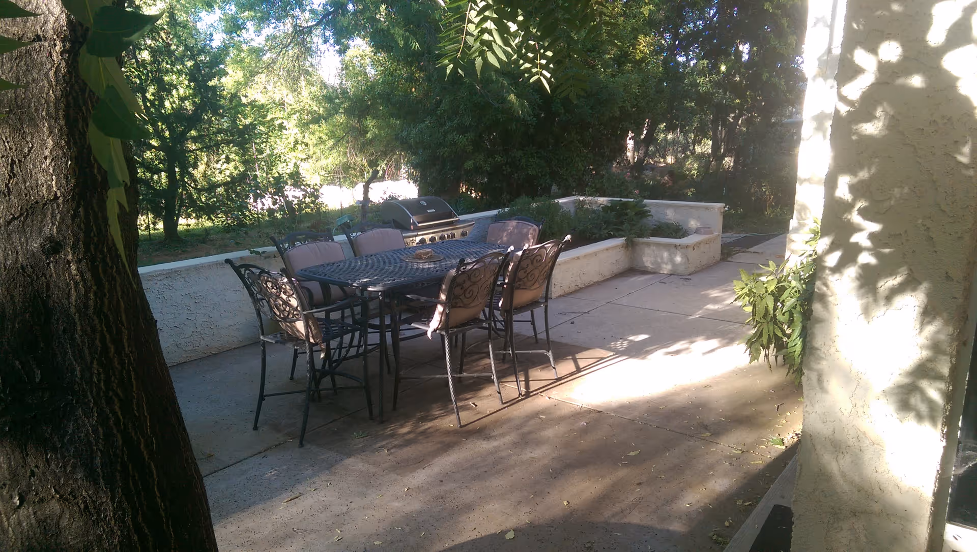 Outdoor patio area with a metal table and six cushioned chairs, a barbecue grill, surrounded by trees and greenery, with sunlight casting shadows on the concrete floor and walls.