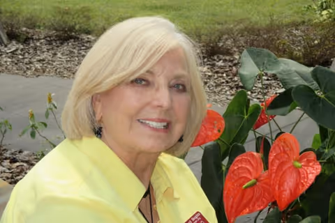 A smiling elderly woman with short blonde hair wearing a yellow shirt is sitting outdoors near red anthurium flowers and green foliage.