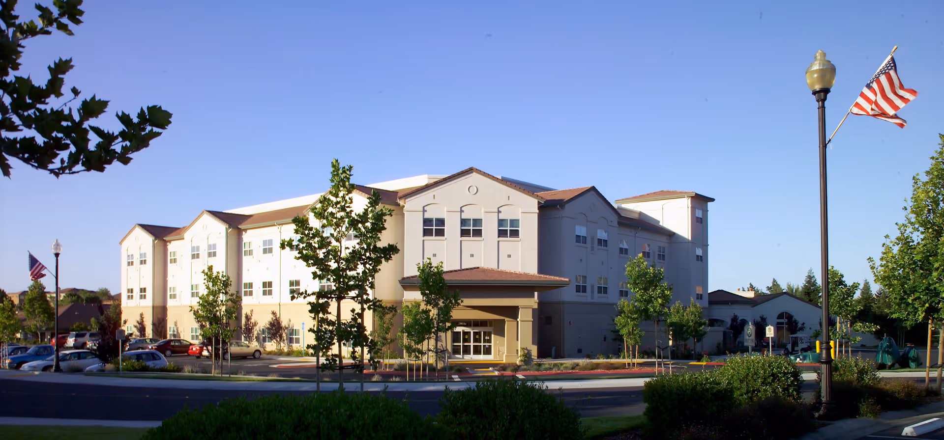 Exterior view of a three-story senior living facility building with beige and light brown walls, multiple windows, and a covered entrance. The building is surrounded by trees, bushes, a parking lot with cars, and an American flag on a lamppost under a clear blue sky.