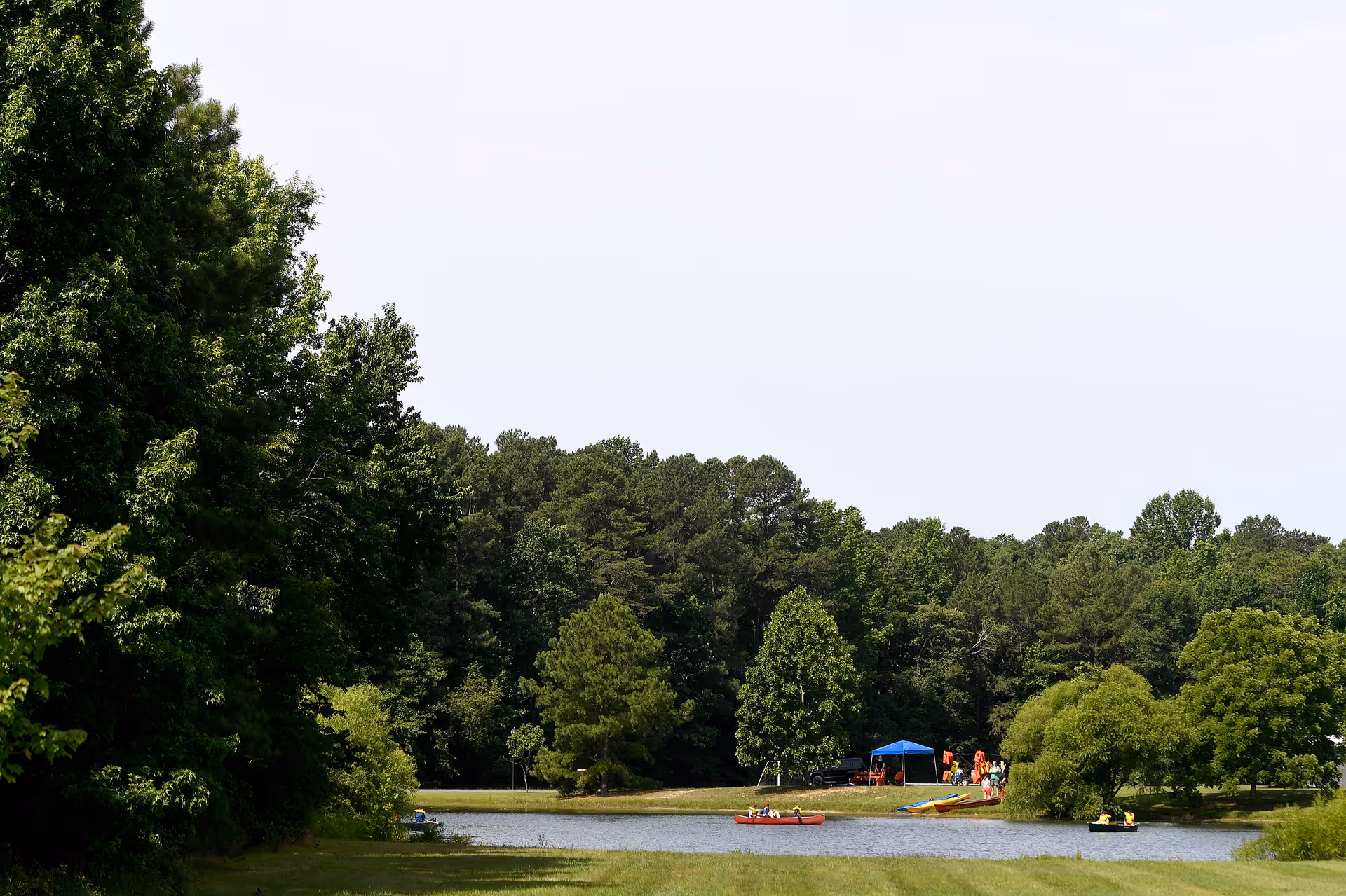 A scenic outdoor area with a small lake surrounded by lush green trees. People are gathered near a blue canopy tent on the far side of the lake, with some individuals in kayaks or canoes on the water.