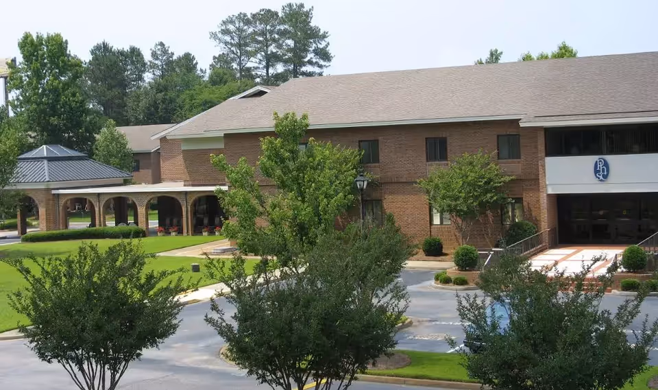 Exterior view of a two-story brick building with a covered entrance and landscaped greenery including trees and bushes in front. The building has a sign with the letters 'BPSC' near the entrance.