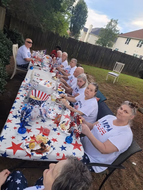 A group of elderly people sitting around a long outdoor table covered with a red, white, and blue star-patterned tablecloth. They are enjoying a meal together in a backyard setting with a wooden fence and houses in the background. The table is decorated with patriotic-themed decorations and food items.