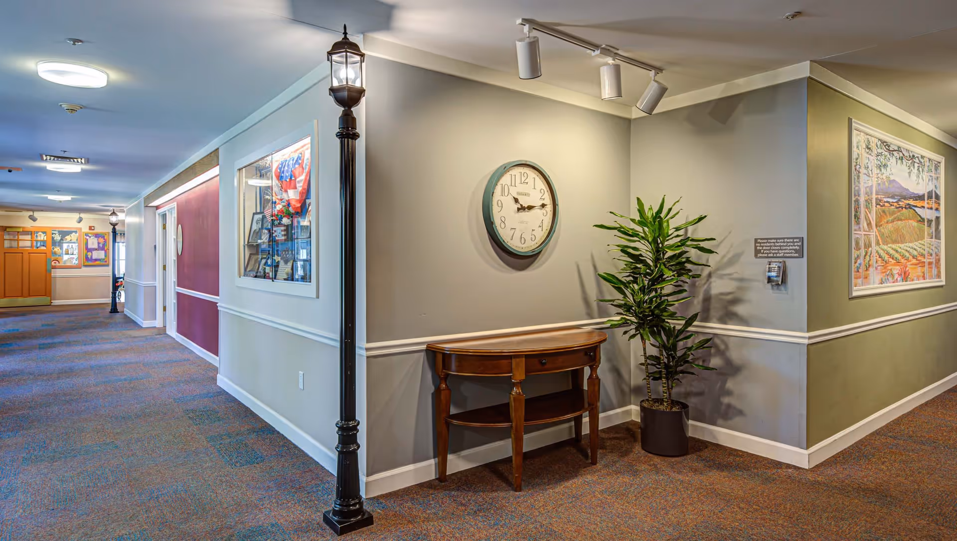 A well-lit hallway in a senior living facility with carpeted floors and walls painted in neutral tones. There is a decorative streetlamp-style light fixture, a wooden console table with a drawer, a potted plant, a round wall clock, and framed artwork on the walls. The hallway extends into the distance with additional lighting and display cases visible.