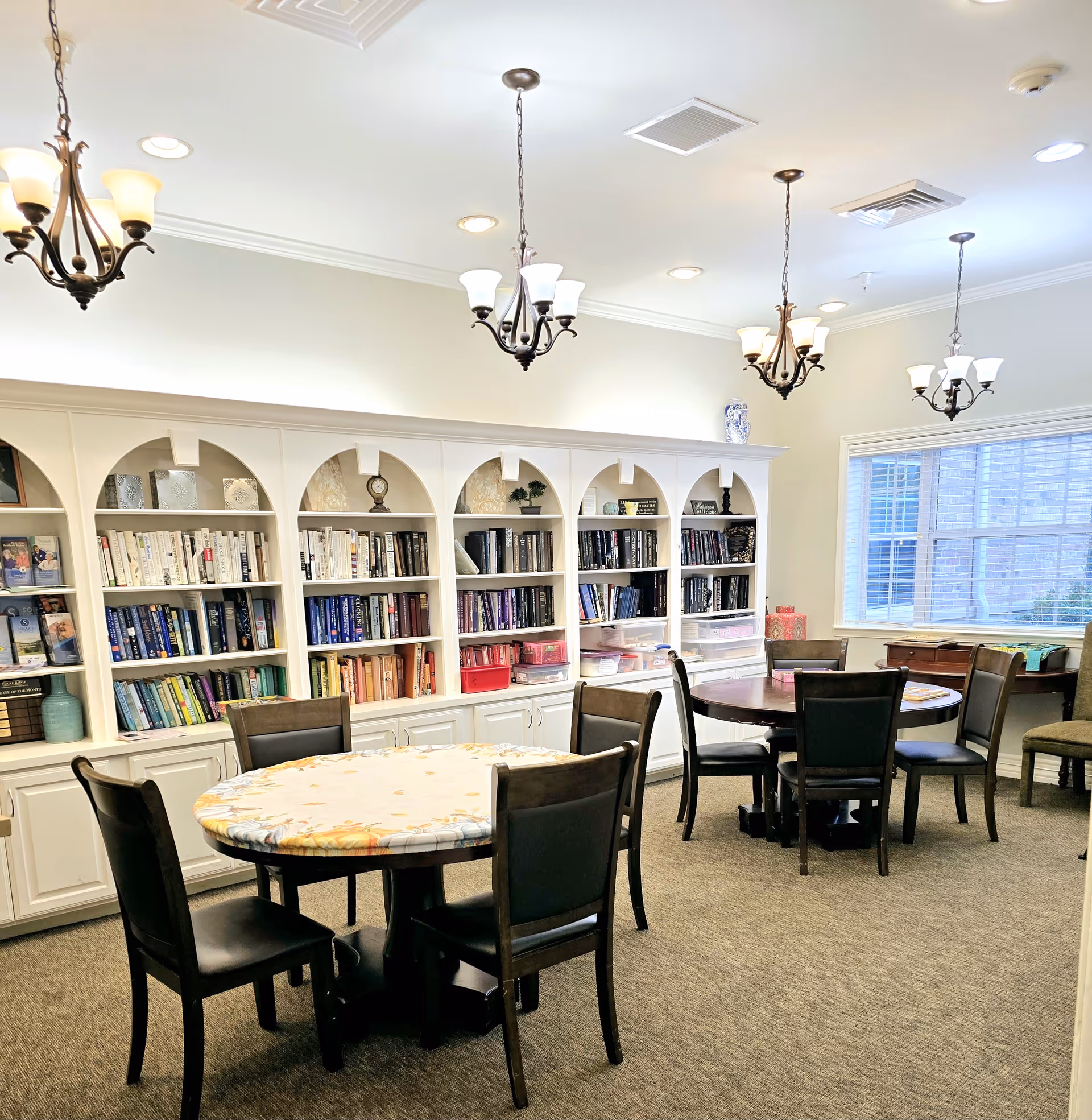 A well-lit room with two round tables covered with tablecloths, each surrounded by four dark wooden chairs. The back wall features a large white built-in bookshelf filled with books and decorative items. Three chandeliers hang from the ceiling, and a large window on the right side lets in natural light.