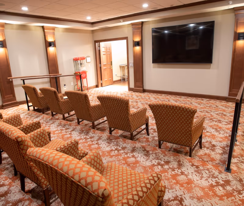 A small theater-style room with patterned orange and beige carpet and matching upholstered chairs arranged in rows facing a large flat-screen TV mounted on the wall. The room has warm wood paneling accents, recessed lighting in the ceiling, and a popcorn machine near an open door leading to another room.