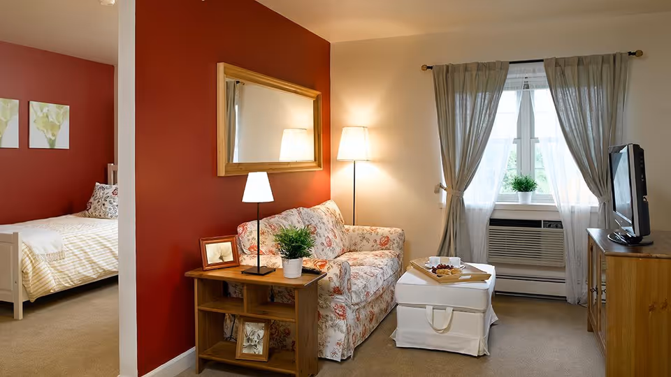 A cozy living room with a floral patterned loveseat, a wooden side table with a lamp and a small plant, a white ottoman with a tray of snacks, and a TV on a wooden cabinet. The room has beige carpet, light-colored walls with one red accent wall, and a window with sheer curtains and an air conditioning unit. A bedroom with a bed and floral pillows is partially visible through an open doorway.