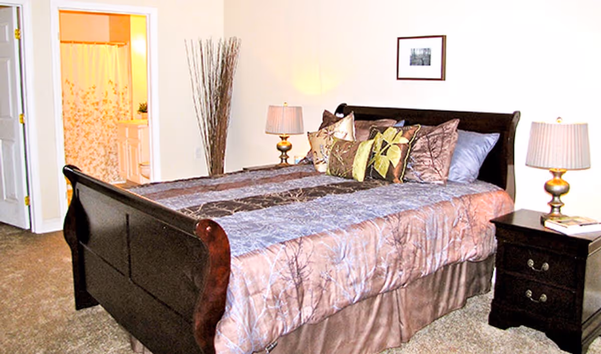 A bedroom with a large wooden bed covered with a patterned comforter and multiple decorative pillows. There are two bedside tables with lamps on each side of the bed. A framed picture hangs on the wall above the bed. An open door reveals a bathroom with a shower curtain and vanity.