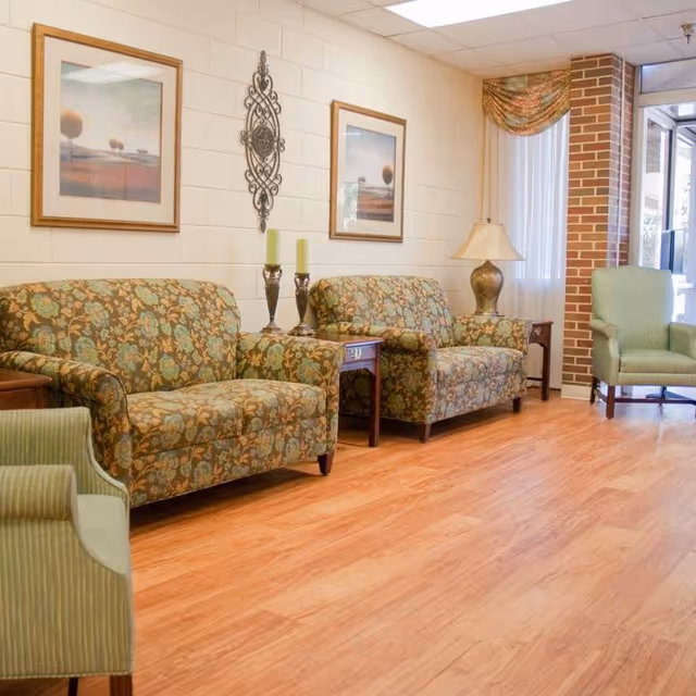 A cozy sitting area in a senior living facility with two floral-patterned loveseats, a green armchair, wooden side tables, decorative candles, a table lamp, framed landscape artwork, and a wall-mounted ornamental piece. The room has light wood flooring, white brick walls, and a window with a floral valance and vertical blinds.