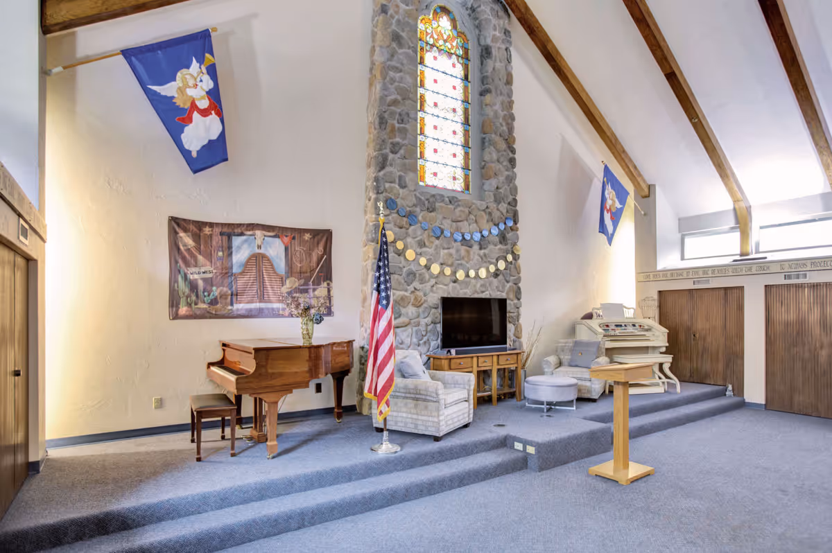 Interior view of a senior living facility room with a stone fireplace featuring a stained glass window above it. The room has a grand piano, an American flag, two armchairs, a small round ottoman, a wooden podium, and a large organ. The ceiling has exposed wooden beams and there are banners with angel designs hanging on the walls.