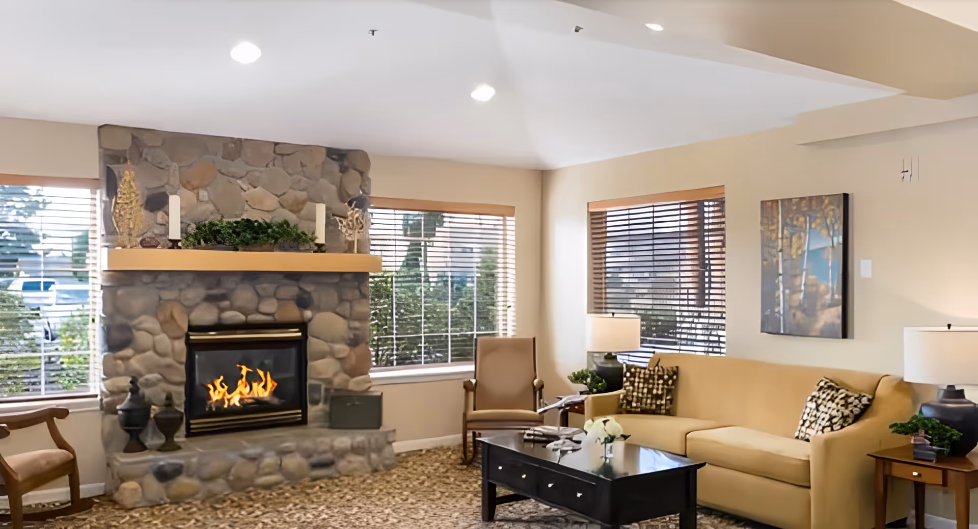 A cozy living room with a stone fireplace featuring a lit fire, two large windows with wooden blinds, a beige sofa with patterned cushions, a wooden armchair, a black coffee table with decorative items, two side tables with lamps, and a painting on the wall.