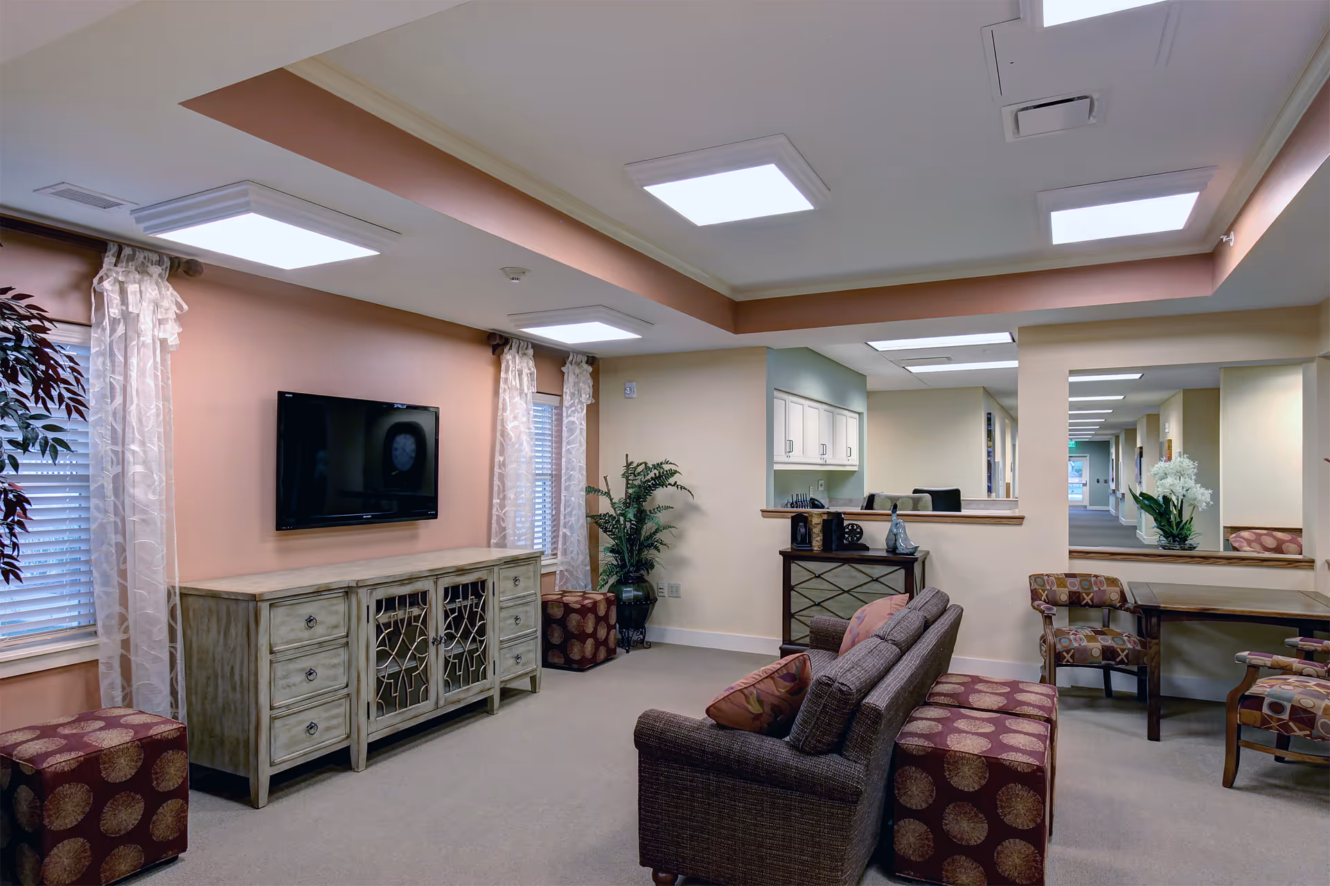 Comfortable common living room with sofas, patterned ottomans, a wall-mounted TV, plants, and a reception counter opening to a hallway.