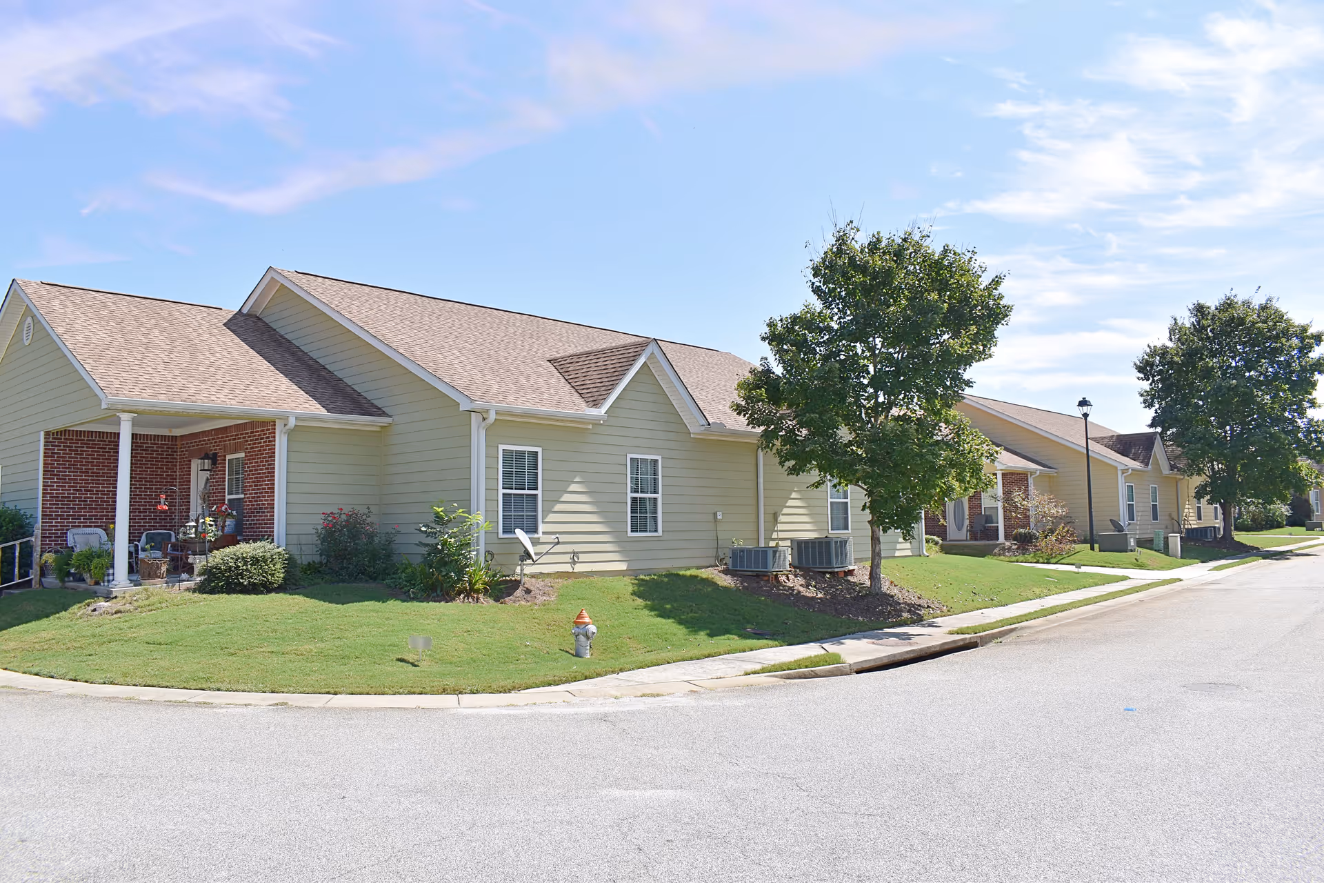 Exterior view of a single-story residential building with light yellow siding and a brown shingled roof. The building has a small covered porch with chairs and plants. There is a well-maintained lawn with bushes and trees along the sidewalk and street under a clear blue sky.