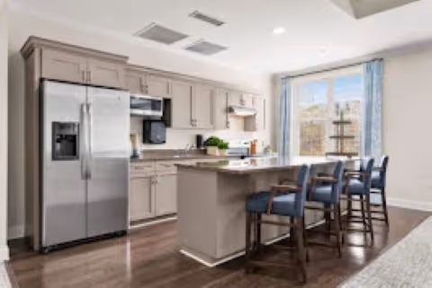 Bright modern kitchen with light gray cabinets, stainless steel refrigerator, built-in microwave, and a kitchen island with four blue cushioned bar stools. Large window with blue curtains lets in natural light.