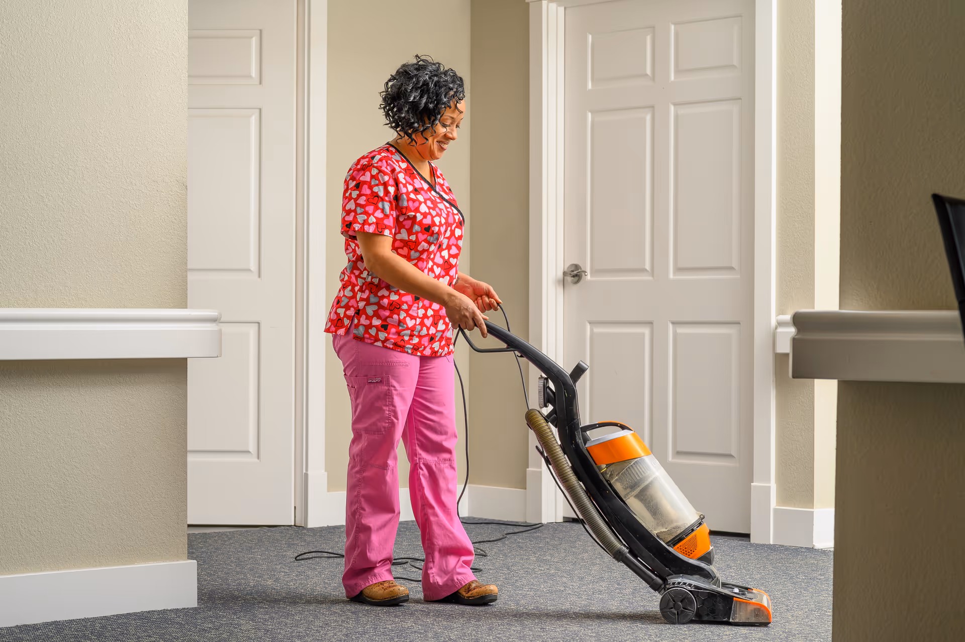 A woman wearing a red shirt with heart patterns and pink pants is vacuuming a carpeted hallway with beige walls and white doors in the background.