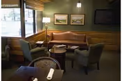A cozy seating area in a senior living facility with a brown leather sofa, two gray armchairs, a round wooden coffee table, and a floor lamp. The walls are green with two framed landscape paintings and a flat-screen TV mounted on the wall. Large windows with blinds allow natural light to enter the room.