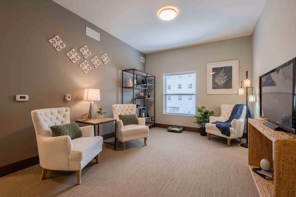 A bright, neutral-toned lounge with three tufted armchairs, side tables, shelving, a TV console and a window.