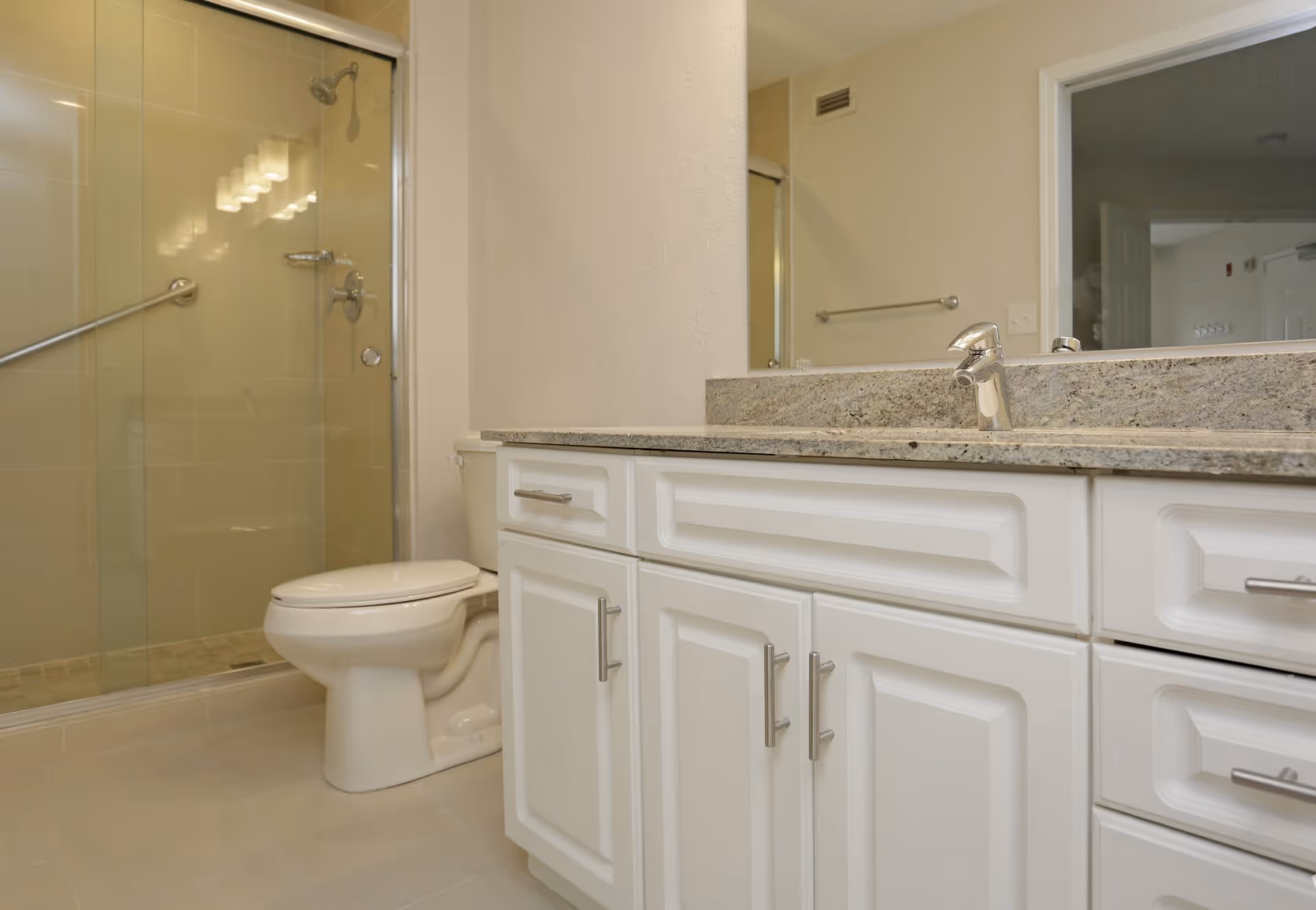 A clean bathroom featuring a white toilet, a glass-enclosed shower with a metal grab bar, and a white vanity with a granite countertop and a single faucet. A large mirror is mounted above the vanity, reflecting part of the room.