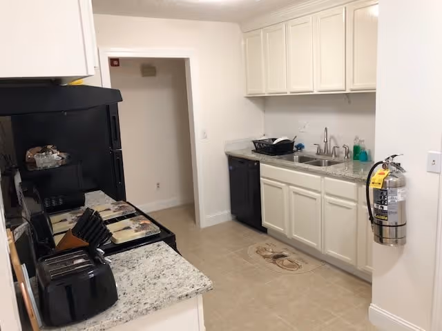 A clean kitchen with white cabinets, granite countertops, a black stove, microwave, toaster, and dishwasher. There is a double sink with a dish rack and soap on the counter. A fire extinguisher is mounted on the wall to the right. The floor is tiled and there is a doorway leading to another room.