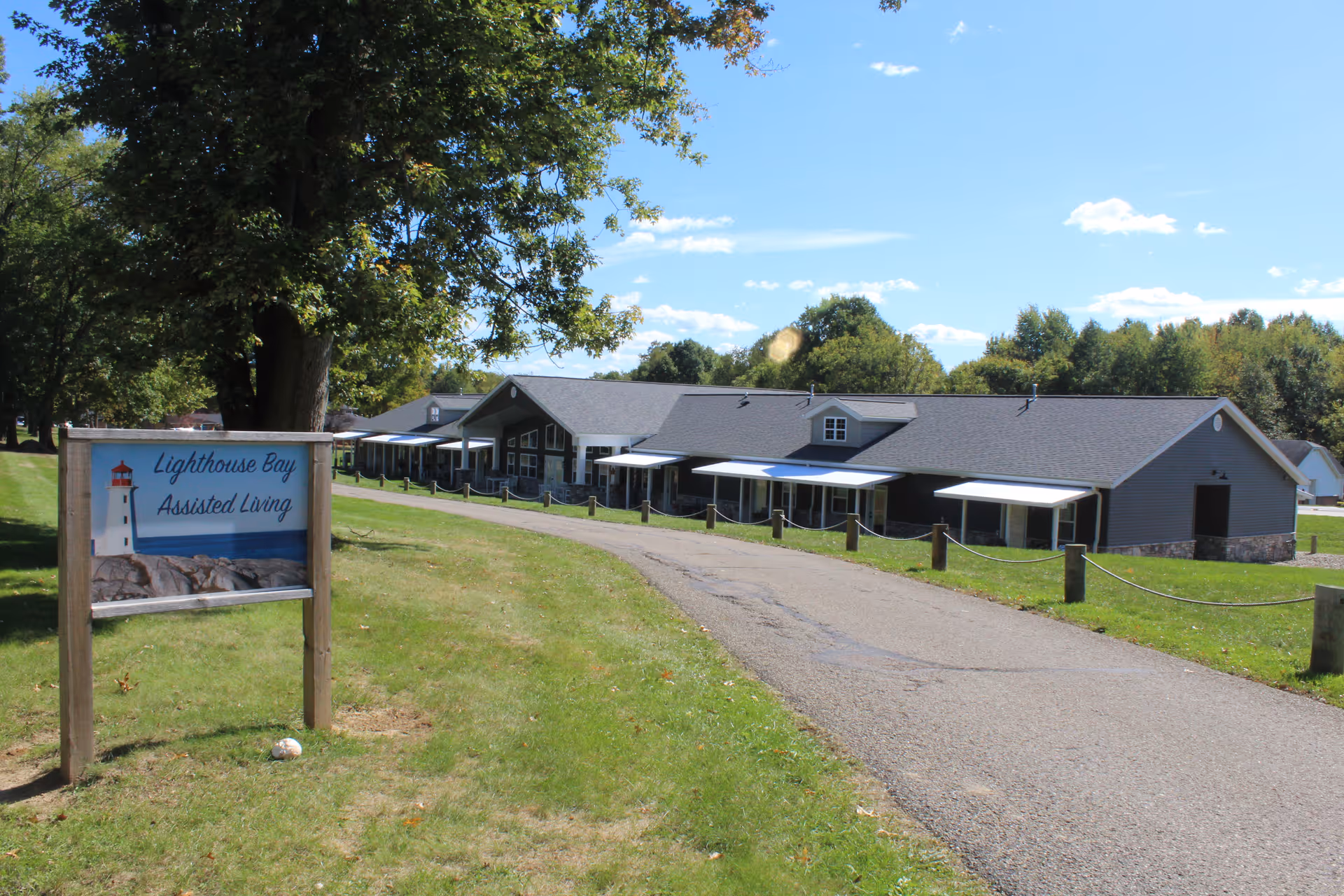 Exterior view of a single-story assisted living facility building with multiple windows and white awnings along a paved driveway. A large tree and green lawn are visible on the left side, along with a sign that reads 'Lighthouse Bay Assisted Living' featuring an image of a lighthouse on rocks. The sky is clear with a few clouds.