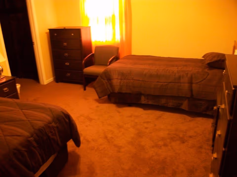 A bedroom with two single beds covered in brown bedspreads, a dark wooden dresser, a matching chest of drawers, a green armchair near a window with yellow curtains, and brown carpeted floor.