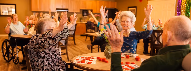 A group of elderly people sitting around tables in a common room playing bingo, raising their hands in celebration. The room has wooden flooring, wooden cabinets, and a warm, inviting atmosphere.