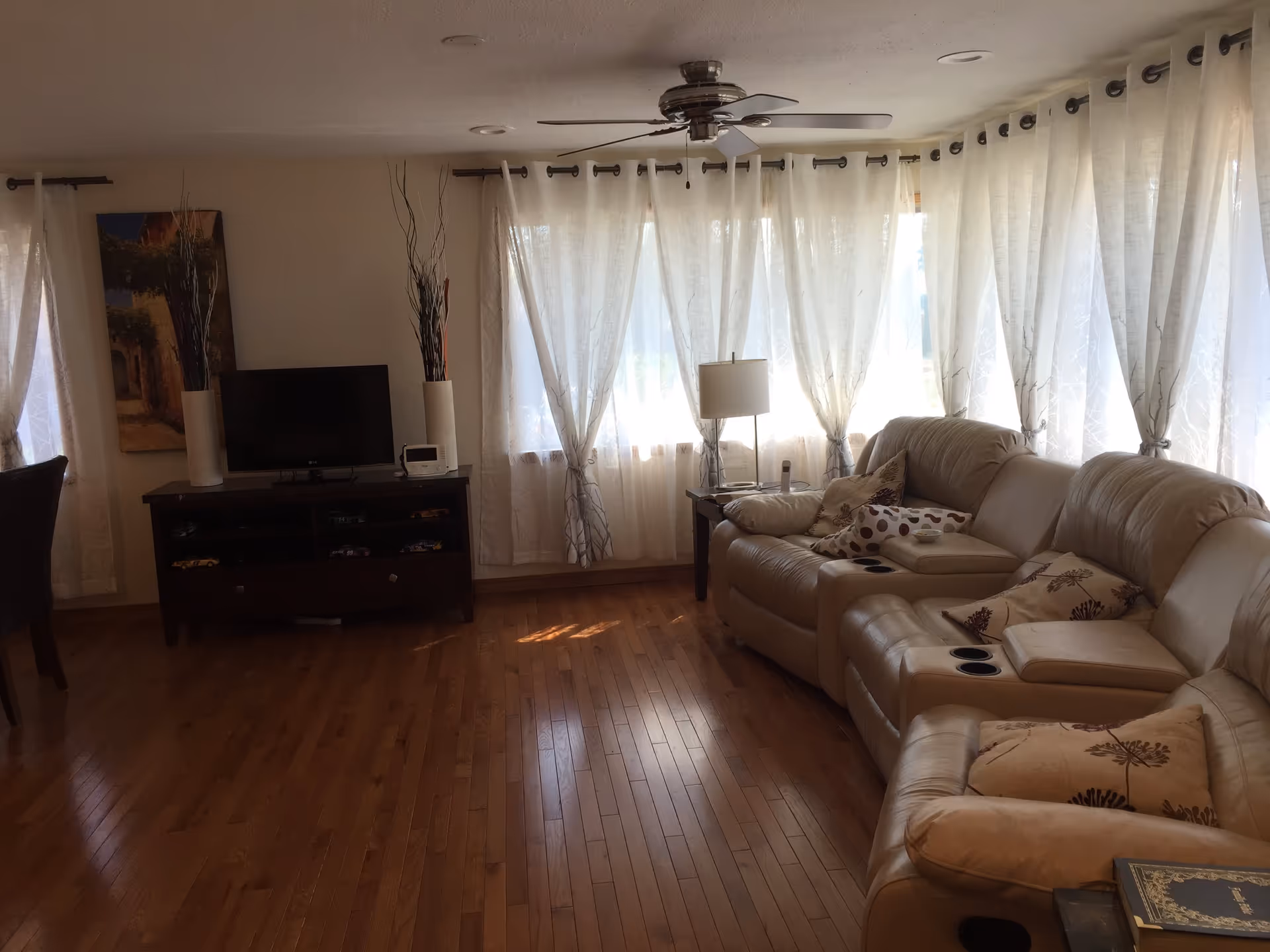 A living room with wooden flooring, a beige leather sectional sofa with patterned cushions, a side table with a lamp, a TV on a dark wooden stand, two tall vases with decorative branches, and large windows covered with sheer white curtains.