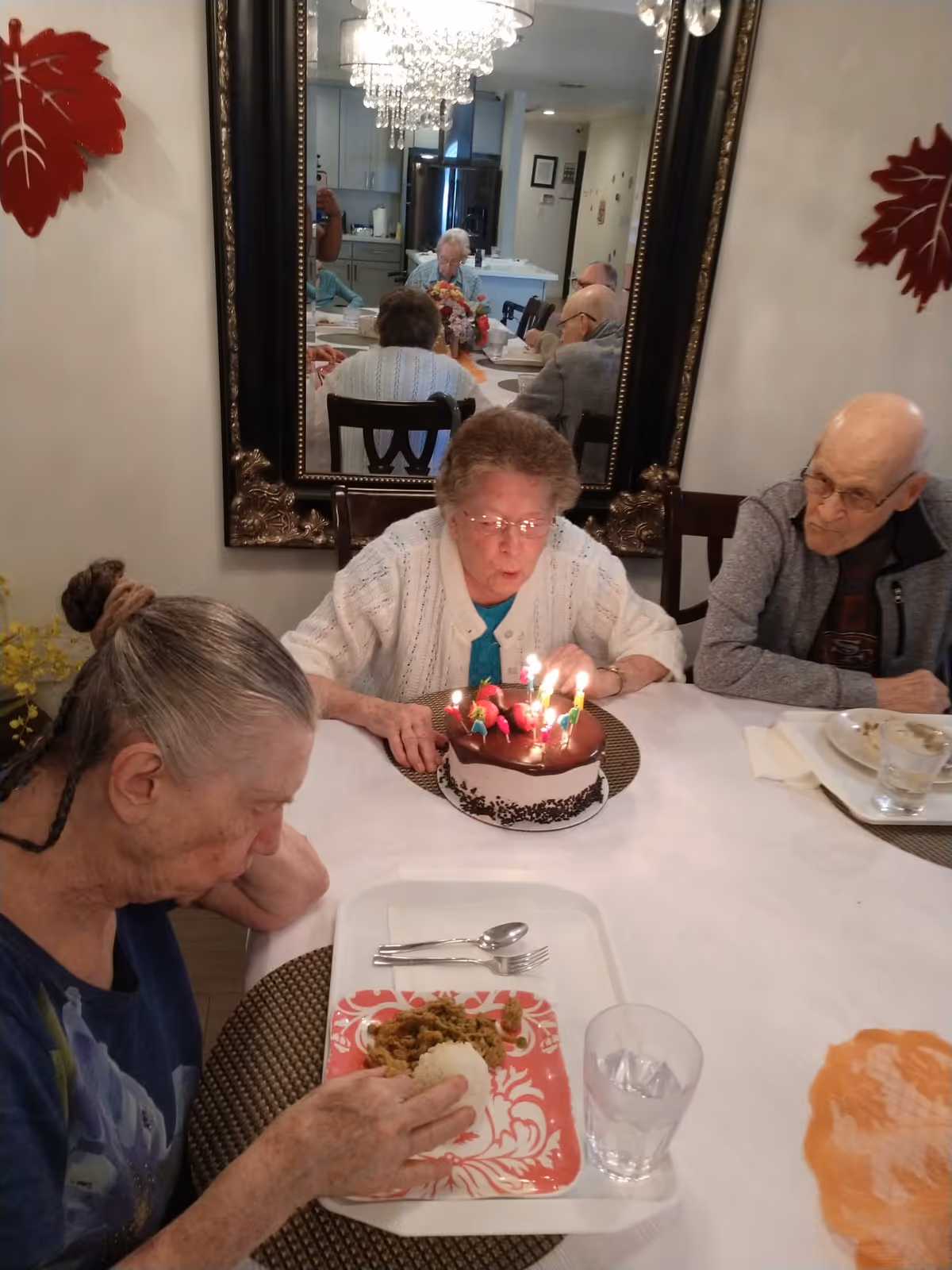 Three elderly people sitting around a dining table in a senior living facility. One woman is blowing out candles on a chocolate birthday cake, while another woman is eating a meal from a tray. A man is sitting next to the woman with the cake, watching. A large mirror on the wall reflects more people sitting at the table in the background. The room is decorated with red leaf wall hangings and a chandelier is visible in the reflection.