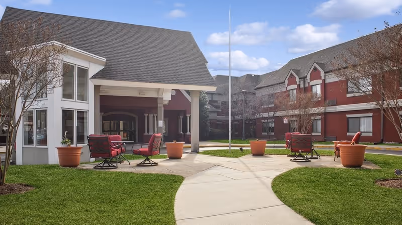 Outdoor courtyard area of a senior living community with red cushioned chairs arranged around small tables on a concrete patio. The courtyard is surrounded by two-story buildings with red and white exterior walls and large windows. There are large potted plants and leafless trees, with a clear blue sky and scattered clouds above.