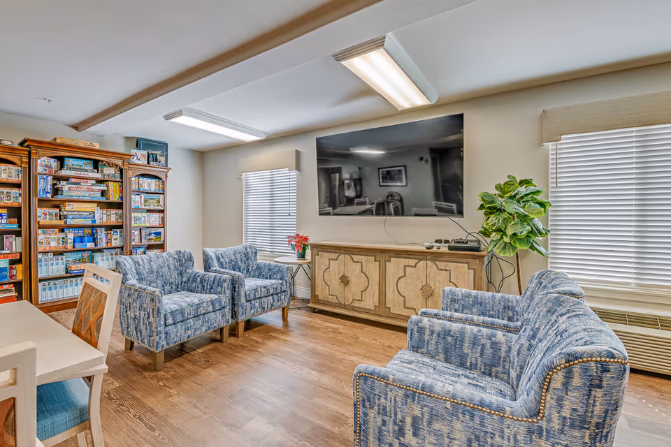 A cozy common area with four blue patterned armchairs arranged around a large flat-screen TV mounted on the wall above a decorative wooden cabinet. There is a tall bookshelf filled with board games on the left side, two windows with blinds, a small round table with a plant, and a wooden floor.