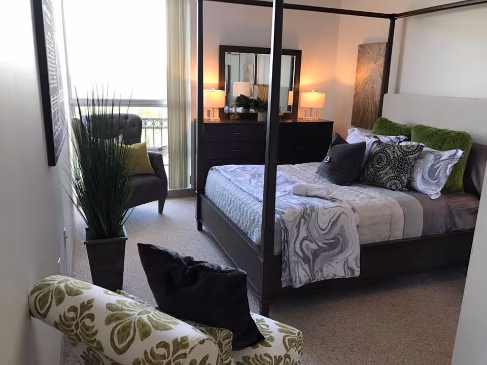 Furnished bedroom featuring a four-poster bed with decorative pillows, a dresser with lamps, and seating by a window.