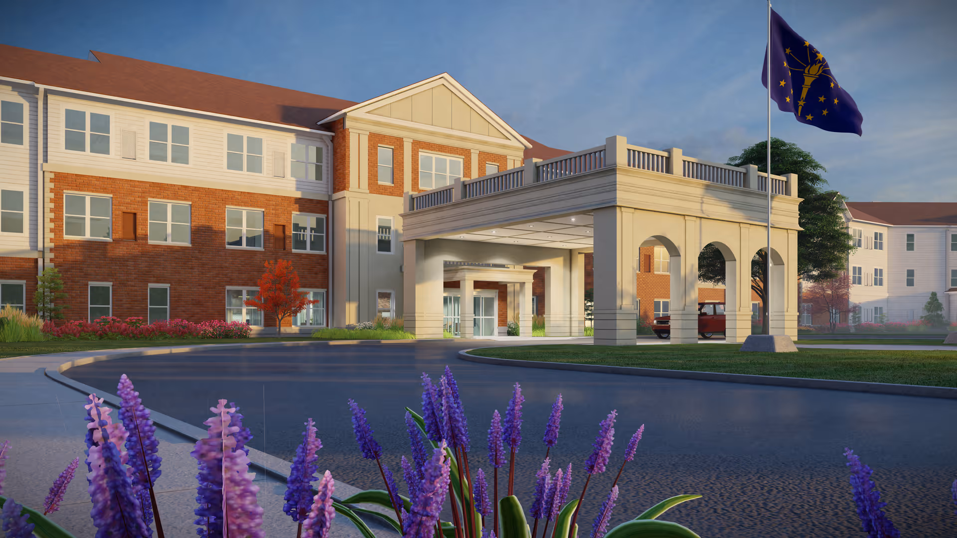 Exterior view of a senior living facility named Harmony at Elkhart, showing a large three-story building with a covered entrance, landscaped grounds with purple flowers in the foreground, and a flagpole with a blue flag featuring a yellow torch and stars.