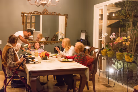A group of elderly women sitting around a dining table inside a warmly lit room, with a man pouring a drink for one of them. The table is set with plates of food and a centerpiece. There is a large ornate mirror on the wall and a glass door leading to an outdoor area with potted plants and string lights.