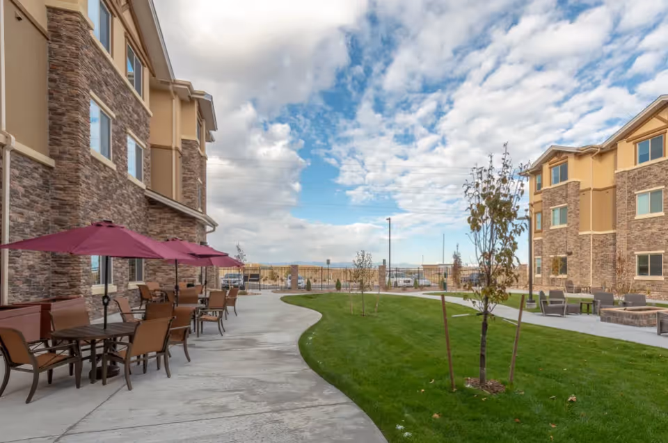 Outdoor courtyard with patio tables and umbrellas, a curved walkway, green lawn, and multi-story stone-faced buildings under a partly cloudy sky.