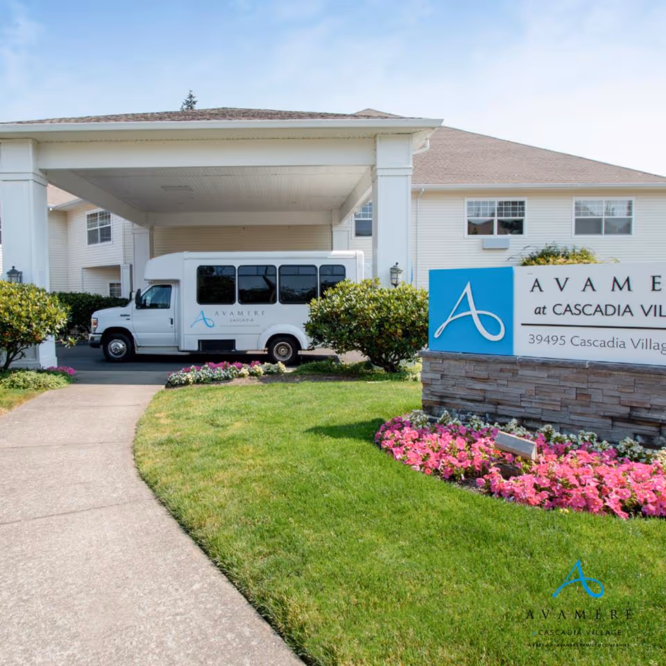 Covered front entrance of Cascadia Village senior living with a shuttle van parked beside a sign and landscaped lawn with flowers.
