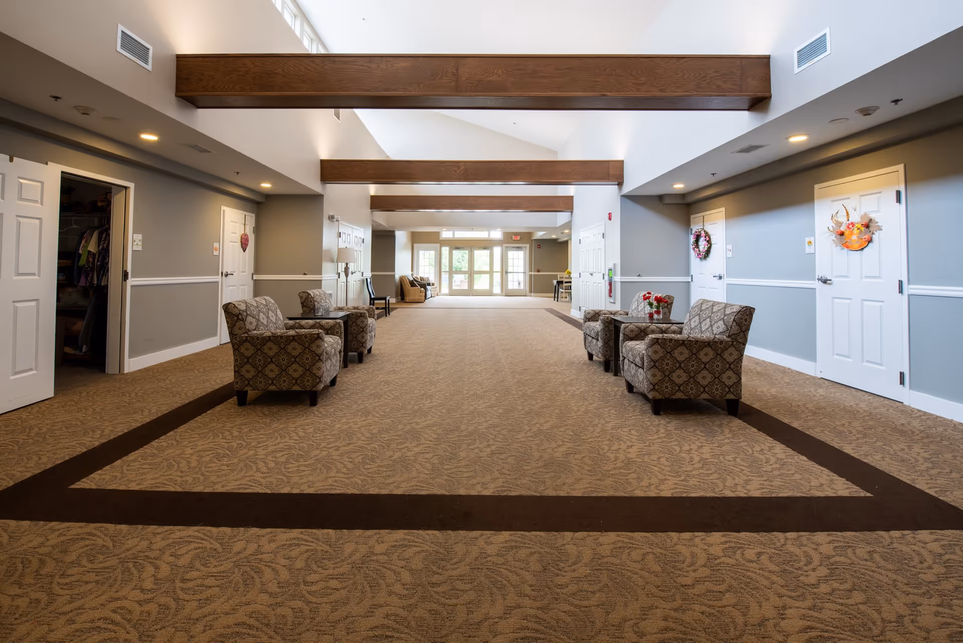 A spacious, well-lit hallway in a senior living facility with patterned carpet and wooden ceiling beams. The hallway features several upholstered armchairs arranged in pairs along the walls, small tables with flower arrangements, and multiple white doors, some decorated with wreaths. Large windows and glass doors at the far end allow natural light to fill the space.
