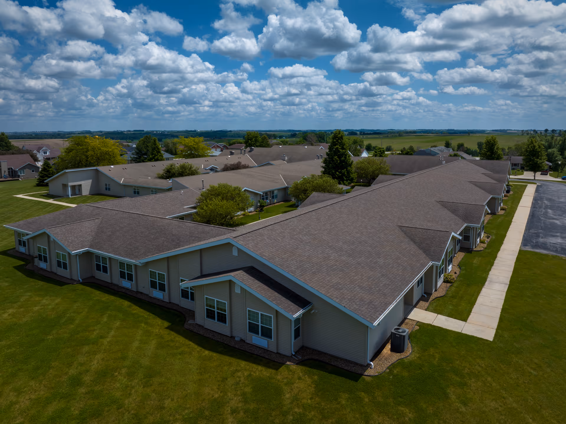 Aerial view of a single-story senior living facility building with multiple wings, surrounded by green lawns and trees under a partly cloudy blue sky.